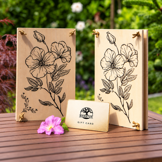 Two rectangular wooden flower presses with black floral line art and brass accessories are displayed on a wooden table outdoors, with a small gift card in front. The background shows blurred green garden foliage.