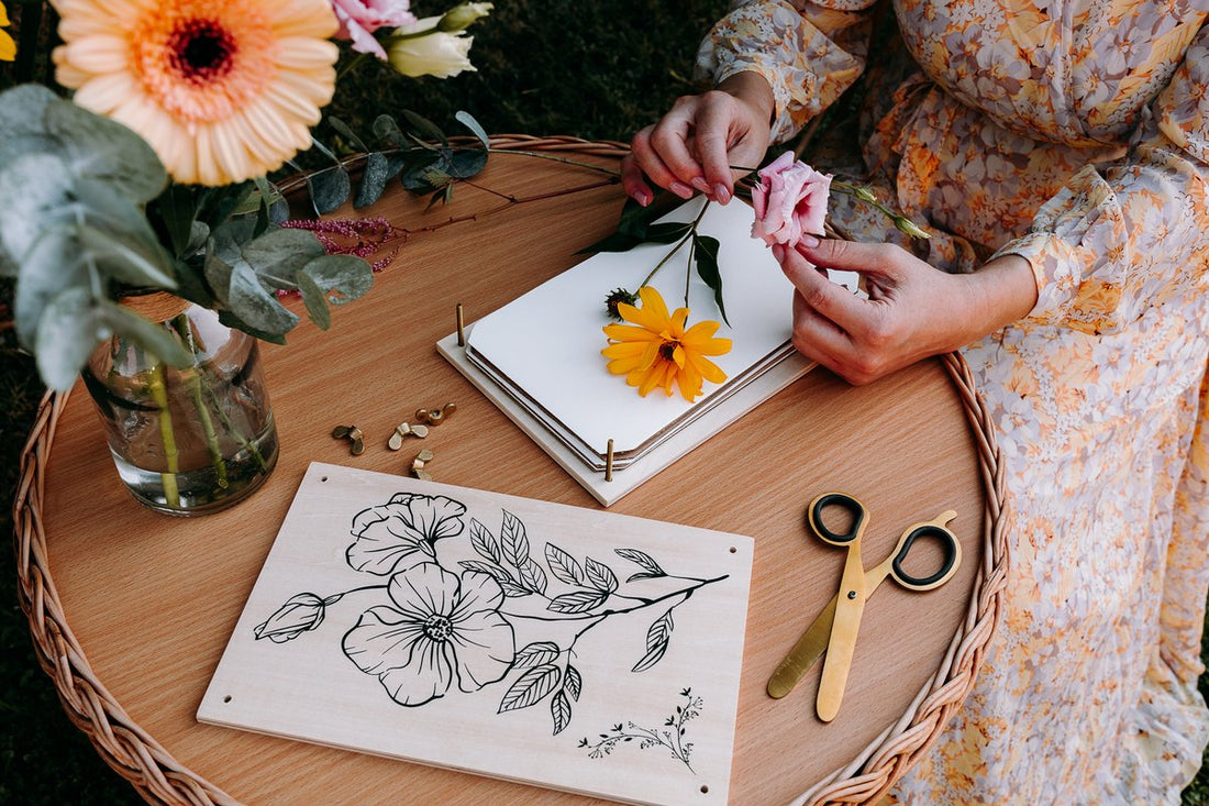 Hands arranging a fresh rose and yellow daisy on an open Berstuk Travel Flower Press at an outdoor table