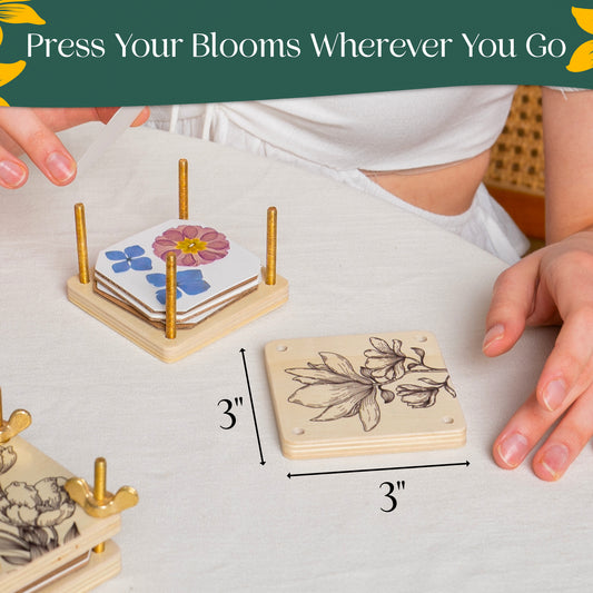 Two wooden flower presses with gold screw posts on a white tablecloth — one holding pressed pink, yellow, and blue flowers, the other engraved with a floral line drawing and 3-inch measurement marks. A banner reads 'Press Your Blooms Wherever You Go
