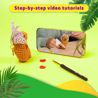 Crocheted capybara watching a phone displaying tutorial, crochet hook, and red heart shapes on a green and yellow background.