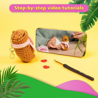 Crocheted capybara toy with a phone showing a tutorial, surrounded by green leaves on a yellow background.