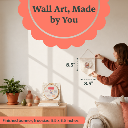 A woman hangs a small fabric wall art piece on a wall in a styled living room. The banner is shown with dimension labels reading 8.5 inches.
