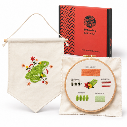 Embroidery starter kit by Berstuk featuring a finished embroidered banner with a green frog and flowers, an embroidery hoop showing stitch guides, and the orange branded kit box on a white background.