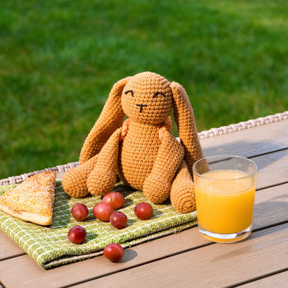Crocheted bunny sat on a wooden table showcasing a mini picnic scene with some orange juice on the side and grapes. 