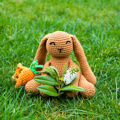 crocheted Bunny sat on grass with a small bunch of flowers and a carrot backpack. 