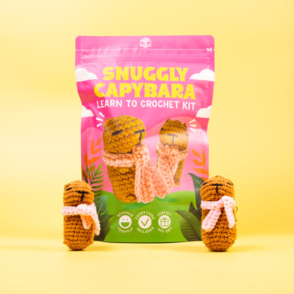 Crocheted capybara toys with a 'Snuggly Capybara' crochet kit packaging on a yellow background