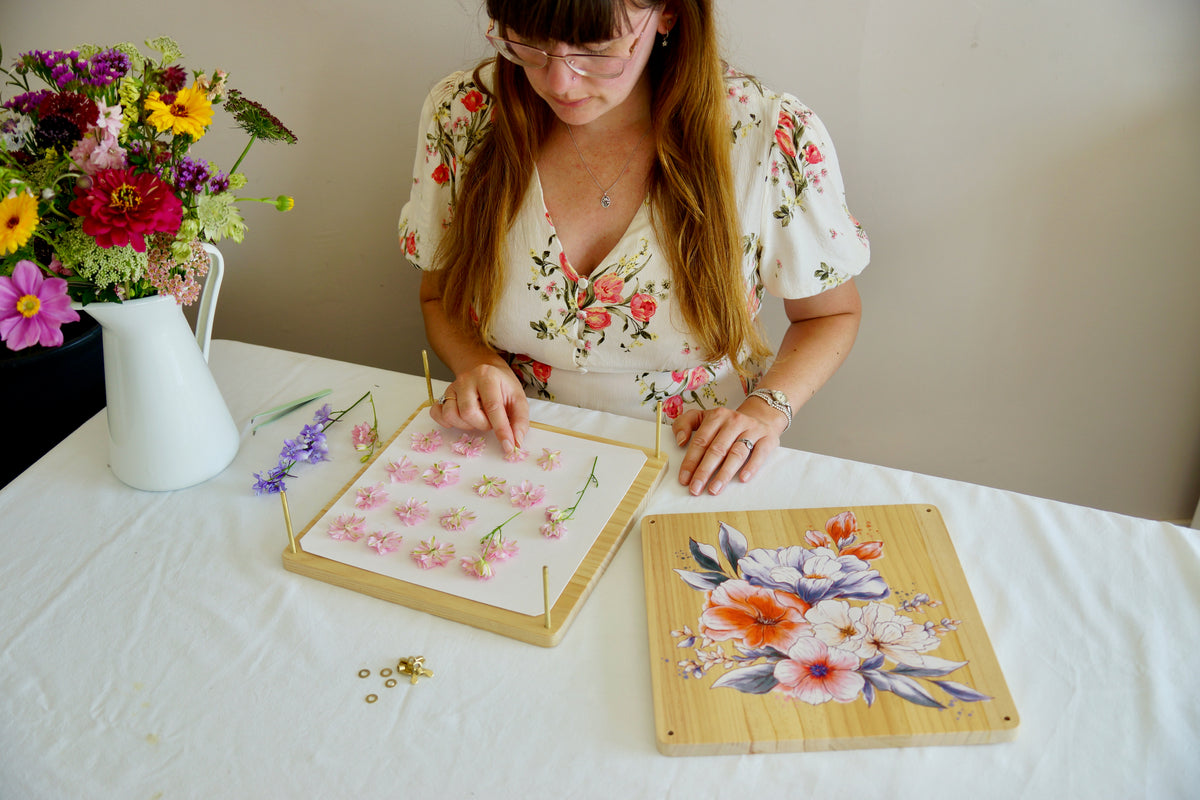 A woman with glasses and long light brown hair sits at a white table, arranging pressed pink flowers on a wooden board. Another wooden board with a colorful floral design is beside her, and a white pitcher of flowers sits on the left.