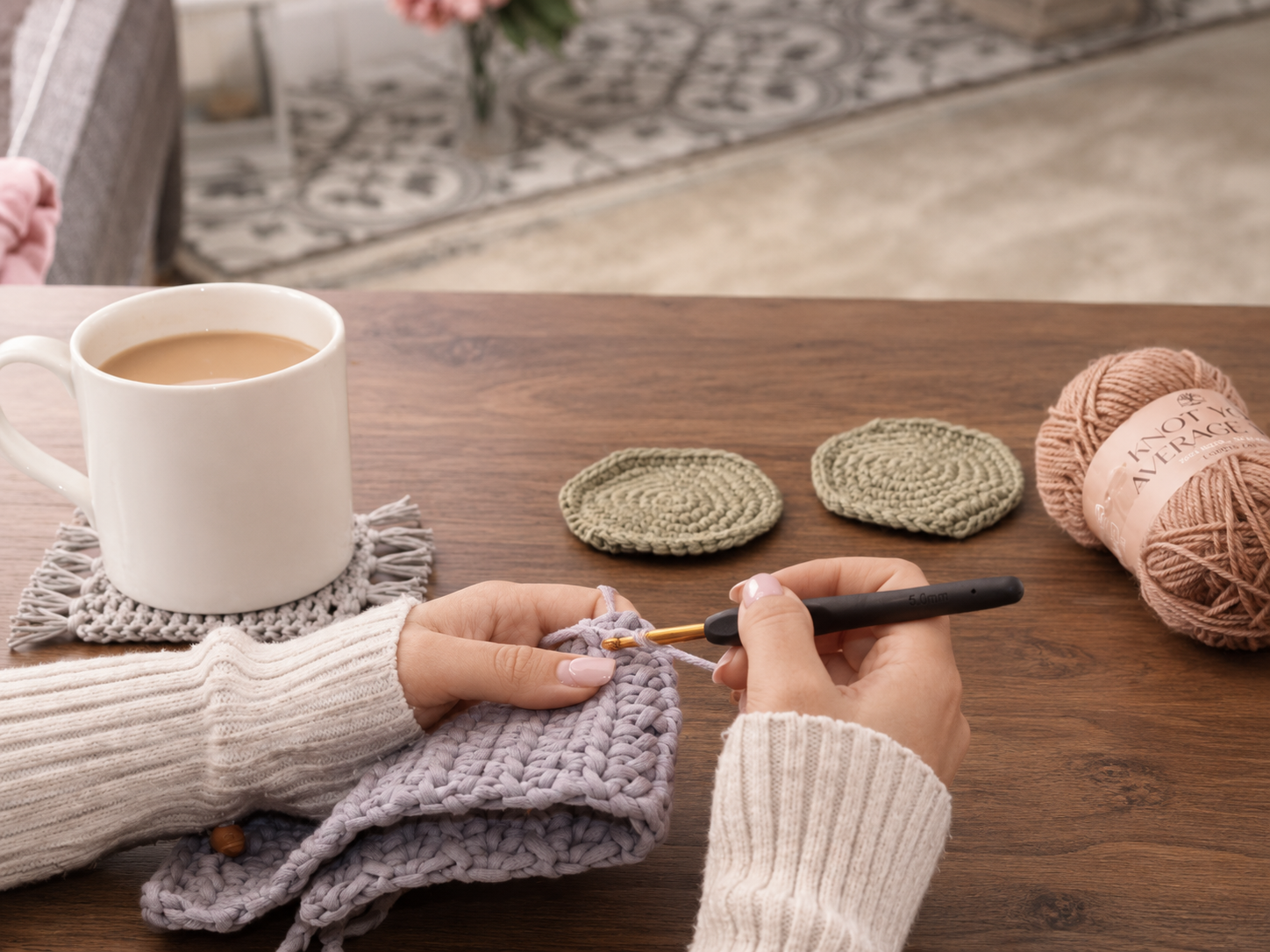 A person crochets a small handmade item on a wooden table, with a white mug on a crocheted coaster, two finished coasters, and yarn balls nearby in a cozy indoor setting.