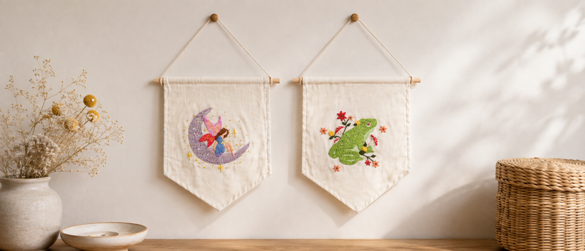 Two cream fabric wall banners hang on a light wall above a wooden surface. The left banner shows a purple crescent moon with a fairy, and the right banner shows a green frog with red flowers.