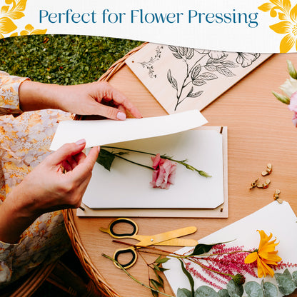 Hands placing a pink flower on blotting paper atop a flower press board, surrounded by scissors and fresh flowers on a wooden table. Text reads 'Perfect for Flower Pressing.